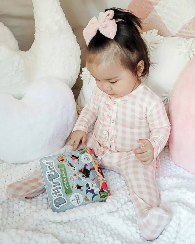A young child in pink plaid pajamas with a pink bow sits on a white knit blanket, holding Ditty Bird UK’s interactive sound book "Animal Songs," surrounded by plush pillows—perfect for early learning and play.