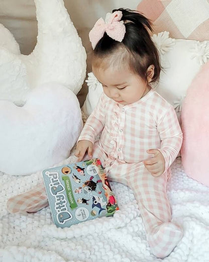 A young child in pink plaid pajamas with a pink bow sits on a white knit blanket, holding Ditty Bird UK’s interactive sound book "Animal Songs," surrounded by plush pillows—perfect for early learning and play.