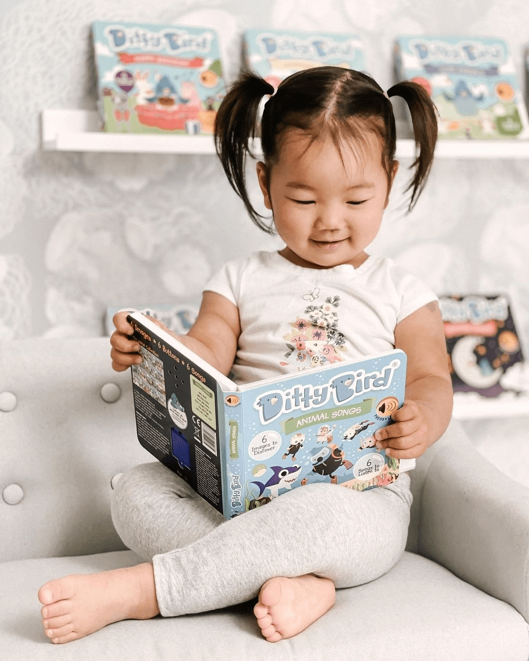 A smiling young child with pigtails sits cross-legged on a chair, enjoying Ditty Bird UK’s Animal Songs book. Colorful children's books are displayed nearby—ideal early learning or first birthday gifts for little ones.