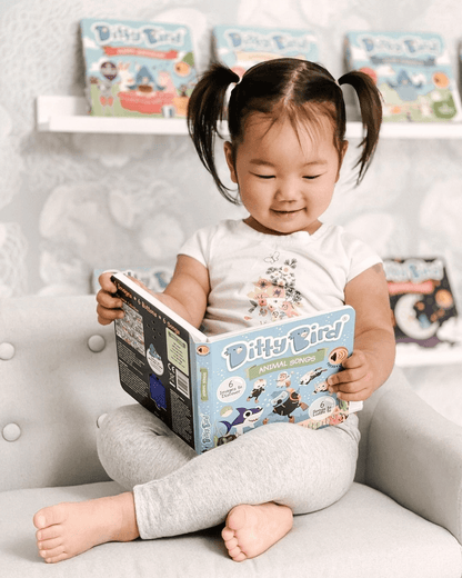 A smiling young child with pigtails sits cross-legged on a chair, enjoying Ditty Bird UK’s Animal Songs book. Colorful children's books are displayed nearby—ideal early learning or first birthday gifts for little ones.