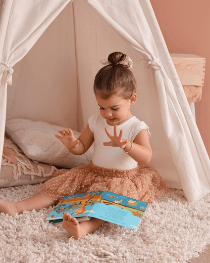 A young girl sits on a soft rug inside a white play tent, wearing a white top and brown tulle skirt. She is looking at Ditty Bird UK’s Animal Songs book—an ideal educational toy or first birthday gift for curious little ones.