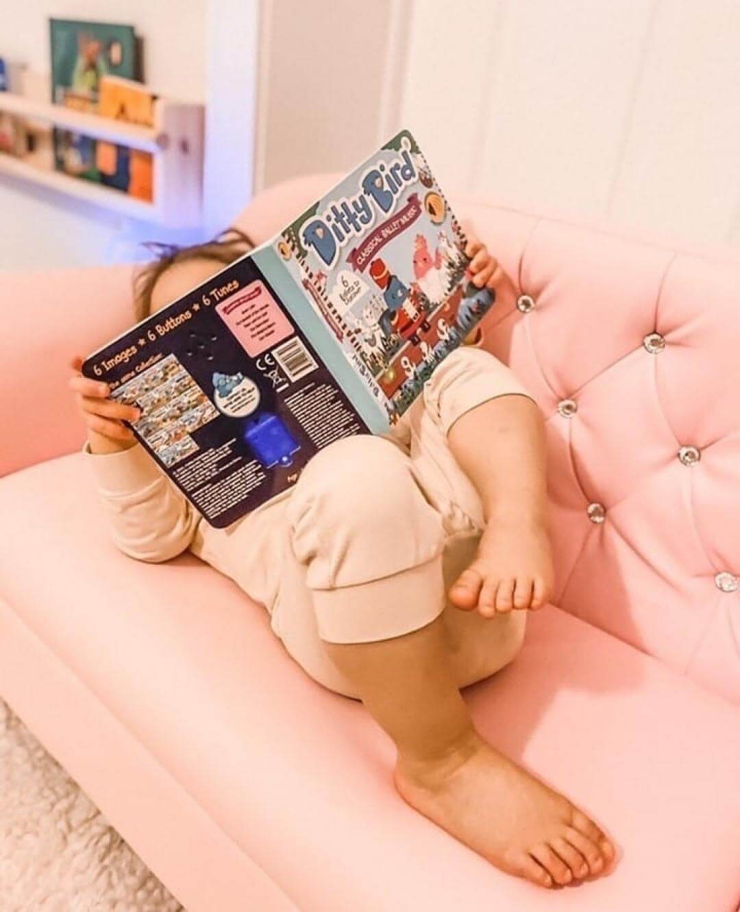 A young child lounges on a light pink, tufted sofa, holding and reading Ditty Bird UK’s "Classical Ballet Music" interactive sound book—one of the best musical toys for toddlers 1-3—with their face mostly hidden and bare feet visible.