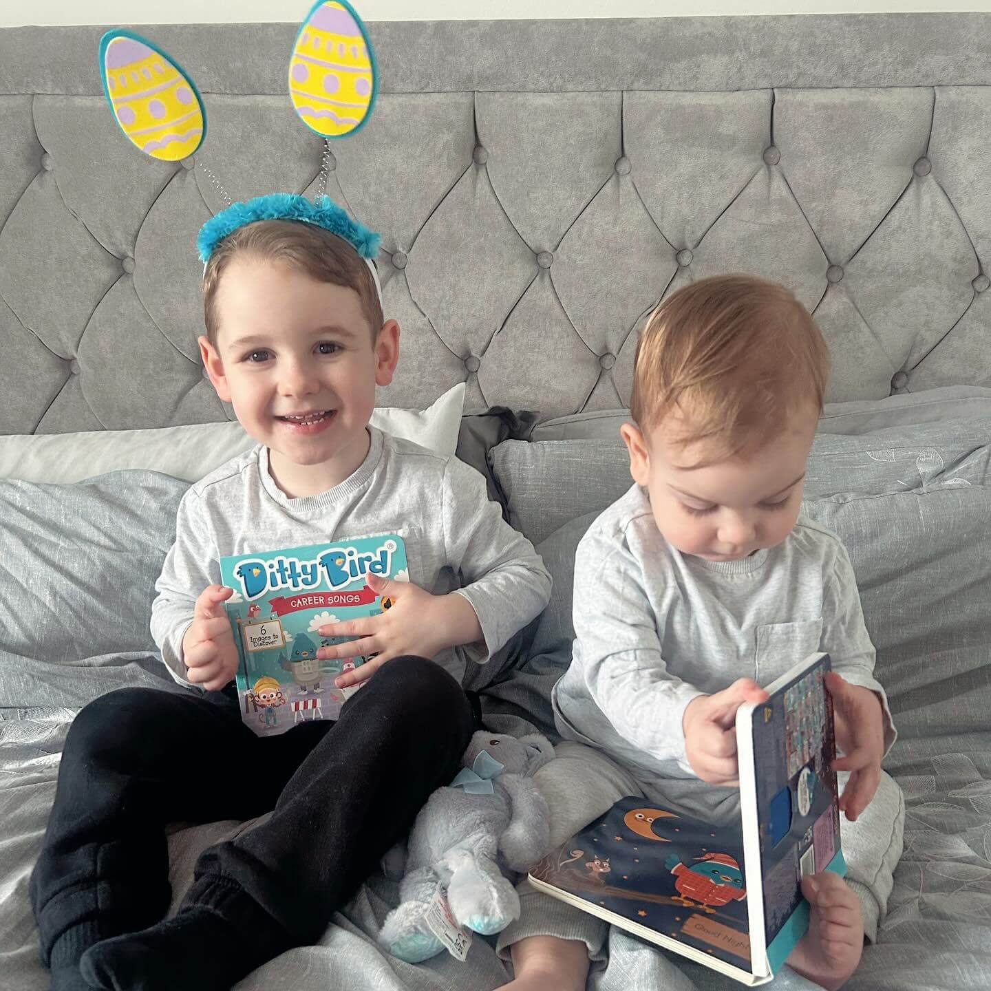 Two young children sit on a bed. The older child, smiling in blue and yellow bunny ears, holds Ditty Bird UK’s interactive sound book “Career Songs.” Both kids wear grey shirts and black pants as the younger one looks down, focused on early learning.