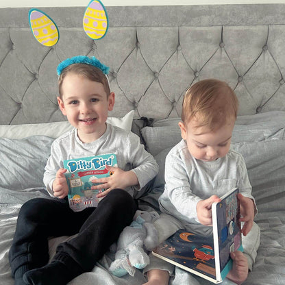 Two young children sit on a bed. The older child, smiling in blue and yellow bunny ears, holds Ditty Bird UK’s interactive sound book “Career Songs.” Both kids wear grey shirts and black pants as the younger one looks down, focused on early learning.