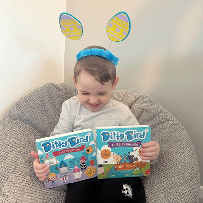 A young child wearing blue and yellow bunny ears smiles on a gray beanbag chair while holding Ditty Bird UK’s Career Songs—a fun interactive sound book, perfect as an educational toy or first birthday gift.