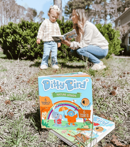 A Ditty Bird UK "Nature Songs" interactive sound book sits on grass, with a woman and young child reading together in the blurred background—an ideal educational toy for outdoor learning.