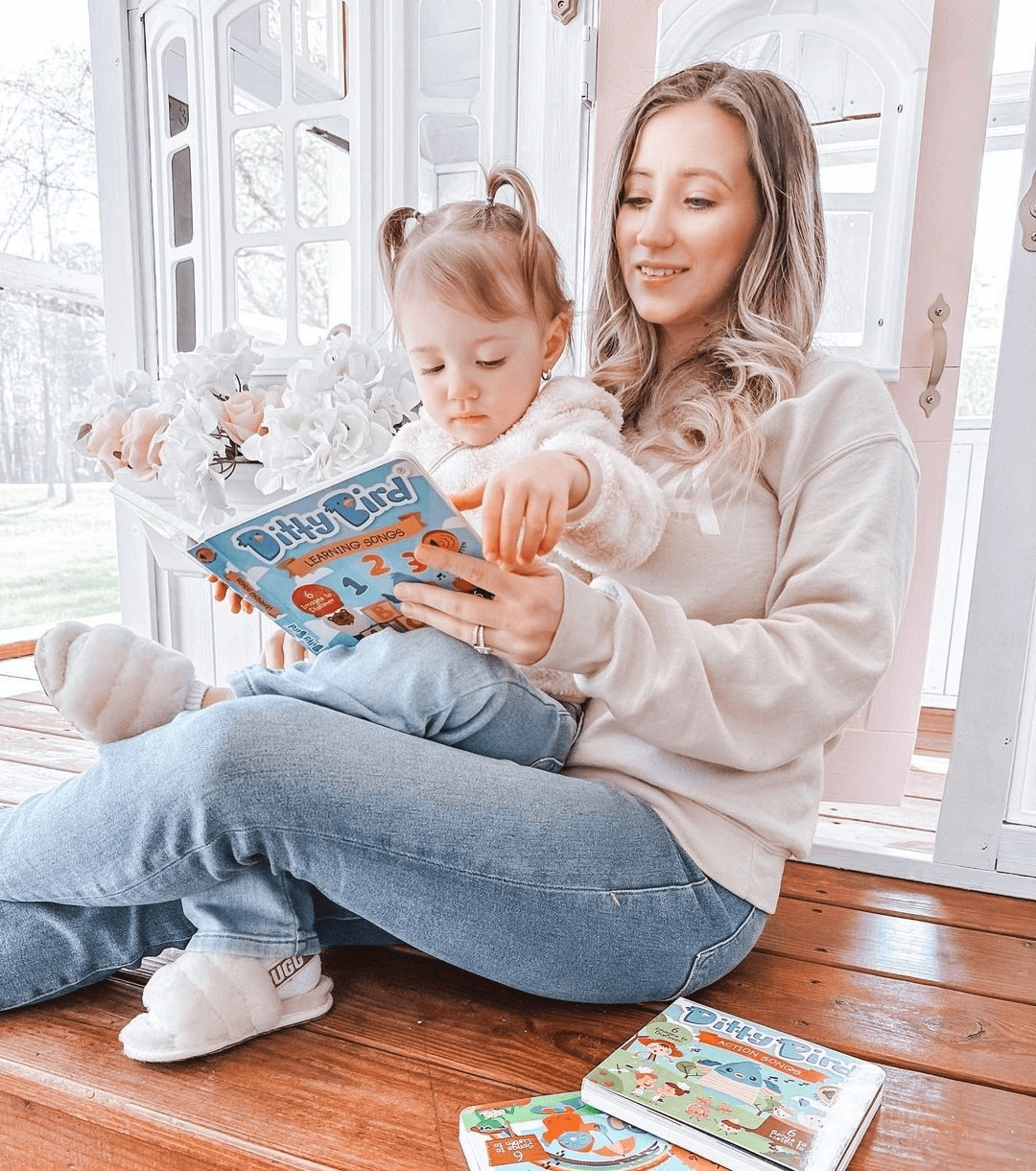 A woman sits on a porch with a young girl on her lap, smiling as they read Ditty Bird UK’s interactive sound book, Nature Songs—an ideal first birthday gift. More children’s books are nearby on the floor.