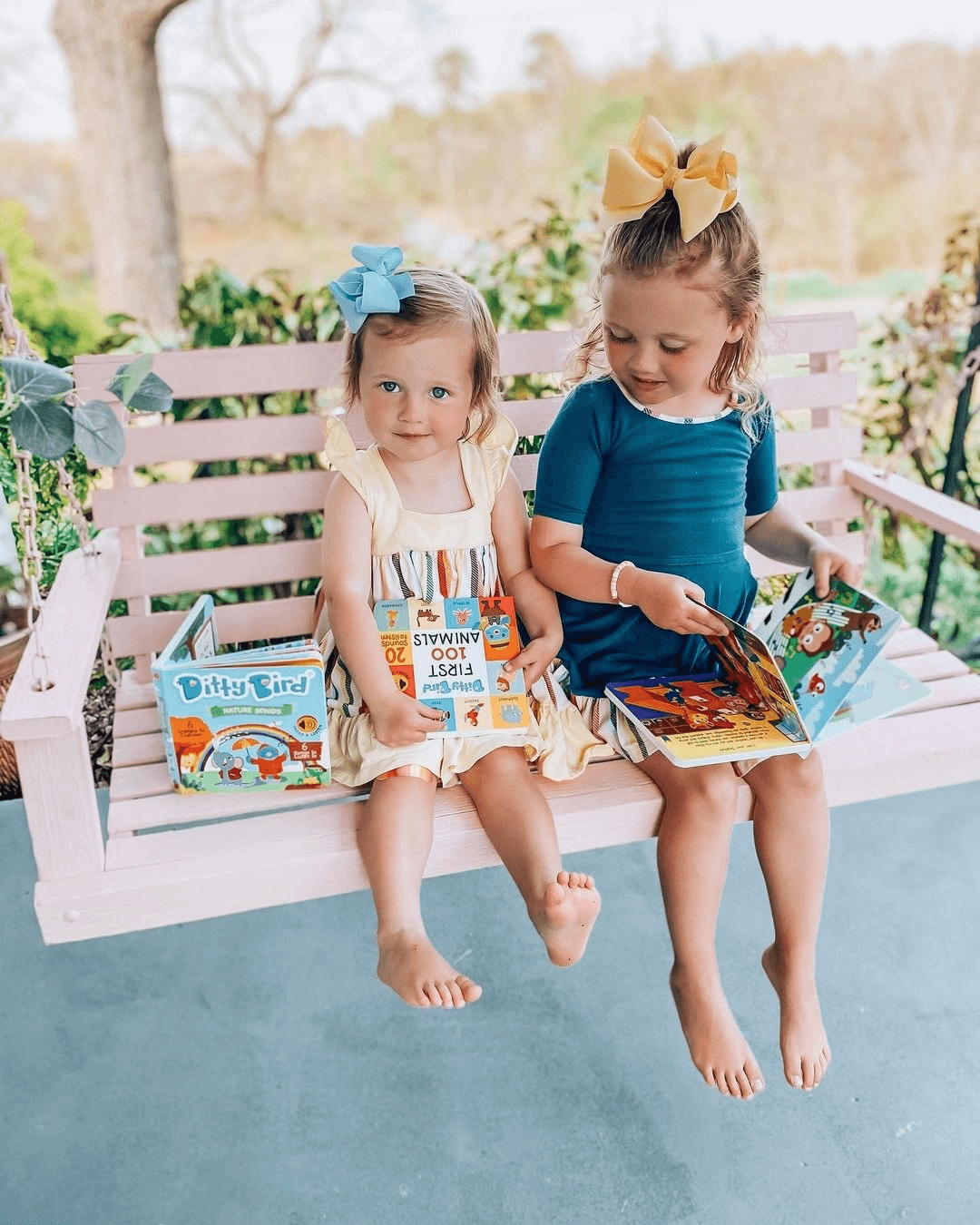 Two young girls with bows in their hair sit barefoot on a pink porch swing, each holding Ditty Bird UK’s "Nature Songs" interactive sound book and smiling, with greenery and a blurred outdoor background behind them.