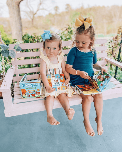 Two young girls with bows in their hair sit barefoot on a pink porch swing, each holding Ditty Bird UK’s "Nature Songs" interactive sound book and smiling, with greenery and a blurred outdoor background behind them.