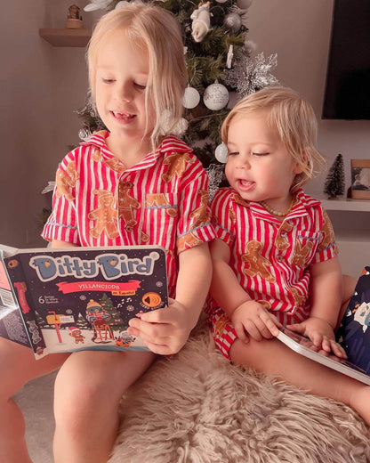 Two young children in matching pajamas sit by a Christmas tree, smiling as they enjoy Ditty Bird UK’s Villancicos en Español—a fun interactive sound book of Spanish Christmas carols, ideal as an educational toy and first birthday gift.