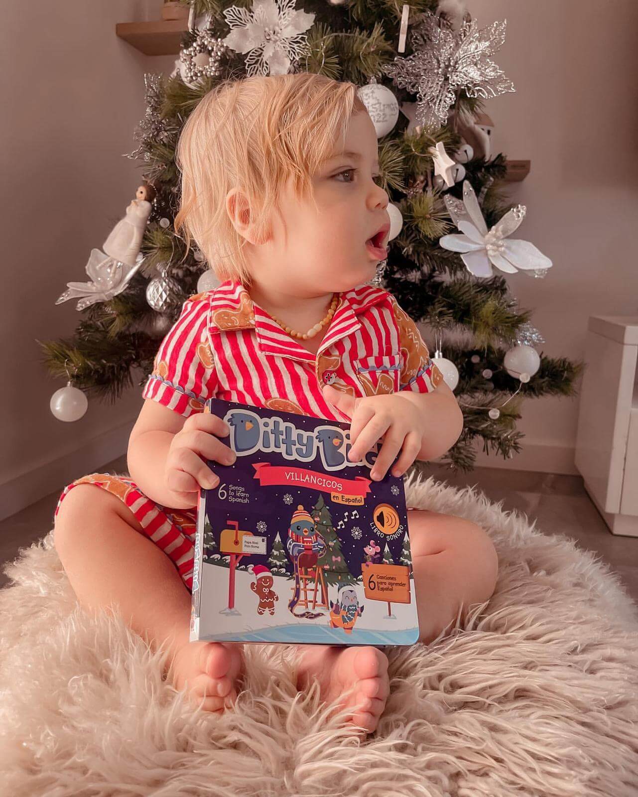A toddler in red-and-white striped pajamas sits on a fuzzy beige rug before a decorated Christmas tree, holding Ditty Bird UK's "Villancicos en Español" book—an ideal educational toy and perfect first birthday gift—while looking to the side.