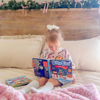 A young girl with a white hair bow sits on a bed with cream pillows and a pink fuzzy blanket, reading the "Villancicos en Español" book by Ditty Bird UK. Garland and string lights decorate the wooden headboard behind her.