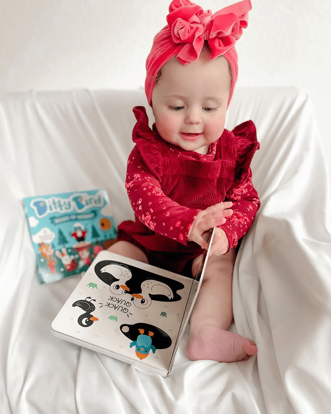 A baby in a bright pink headband and red outfit sits on a white blanket, smiling as they turn a picture book. Ditty Bird UK's "Black & White Animals," one of the best musical toys for toddlers 1–3, is visible in the background.