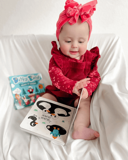 A baby in a bright pink headband and red outfit sits on a white blanket, smiling as they turn a picture book. Ditty Bird UK's "Black & White Animals," one of the best musical toys for toddlers 1–3, is visible in the background.