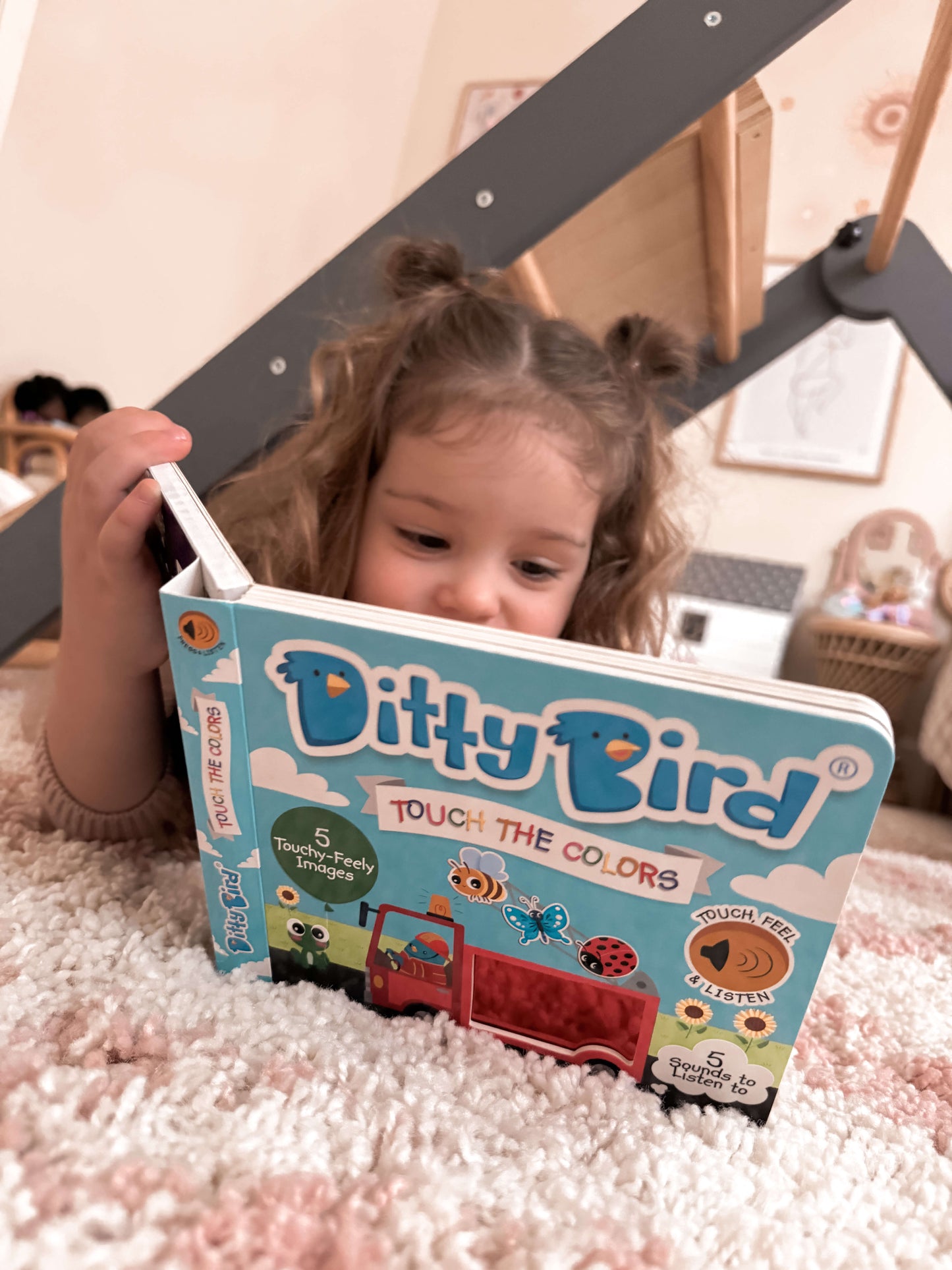 A young child with brown hair in pigtails lies on a soft, patterned rug, intently reading Ditty Bird UK's Touch the Colors interactive sound book—a wonderful educational toy and first birthday gift.