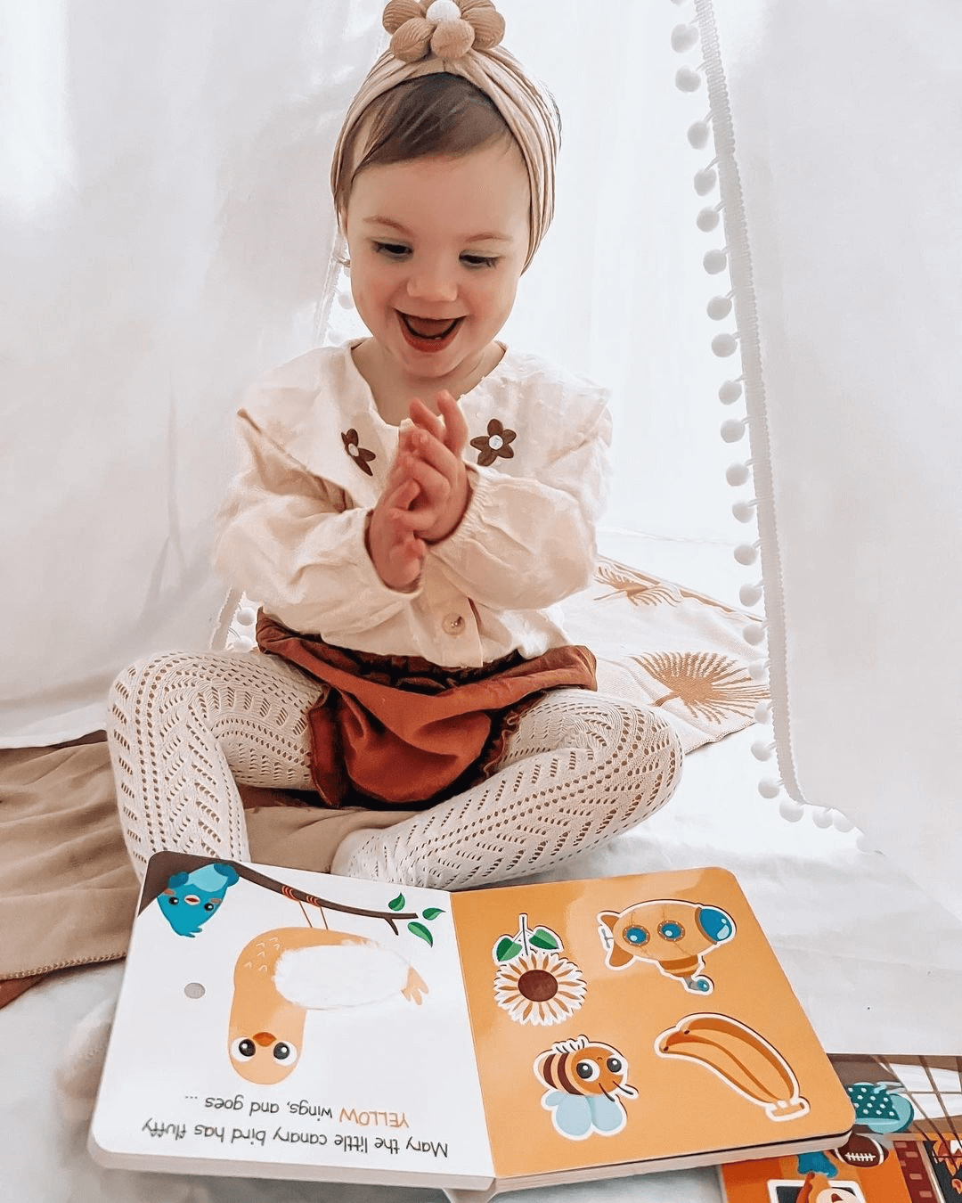 A young child smiles and claps on a soft mat inside a cozy white tent, wearing a headband and patterned tights. In front lies Ditty Bird UK's Touch the Colors, an interactive sound book perfect for early learning fun.