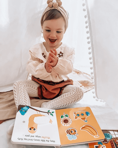 A young child smiles and claps on a soft mat inside a cozy white tent, wearing a headband and patterned tights. In front lies Ditty Bird UK's Touch the Colors, an interactive sound book perfect for early learning fun.