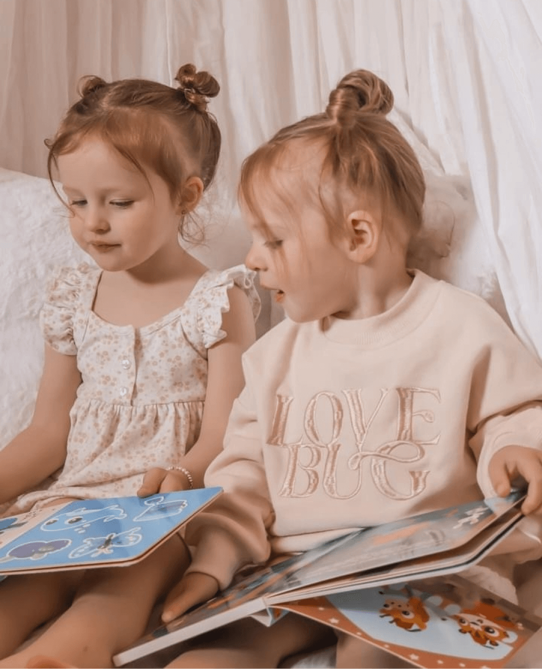 Two young girls with light brown hair in top buns sit together reading the Ditty Bird UK Touch the Colors interactive sound book. One wears a white ruffled dress and the other a light "LOVE BUG" sweatshirt, on a white surface with a soft background.