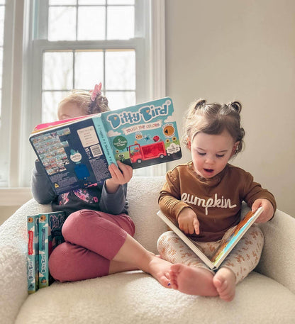 Two young children sit together on a plush chair, each reading. The child on the left holds up Ditty Bird UK’s "Touch the Colors" book, while the child on the right, in a pumpkin sweater, points at pictures in an open book.