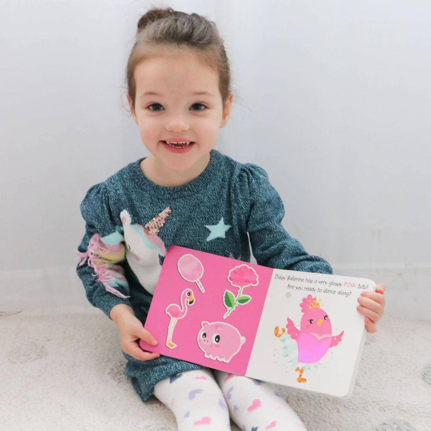 A young girl with brown hair in a ponytail enjoys Ditty Bird UK's Touch the Colors interactive sound book, smiling as she sits on the carpet. Filled with colorful animal illustrations, it's perfect as a first birthday gift.