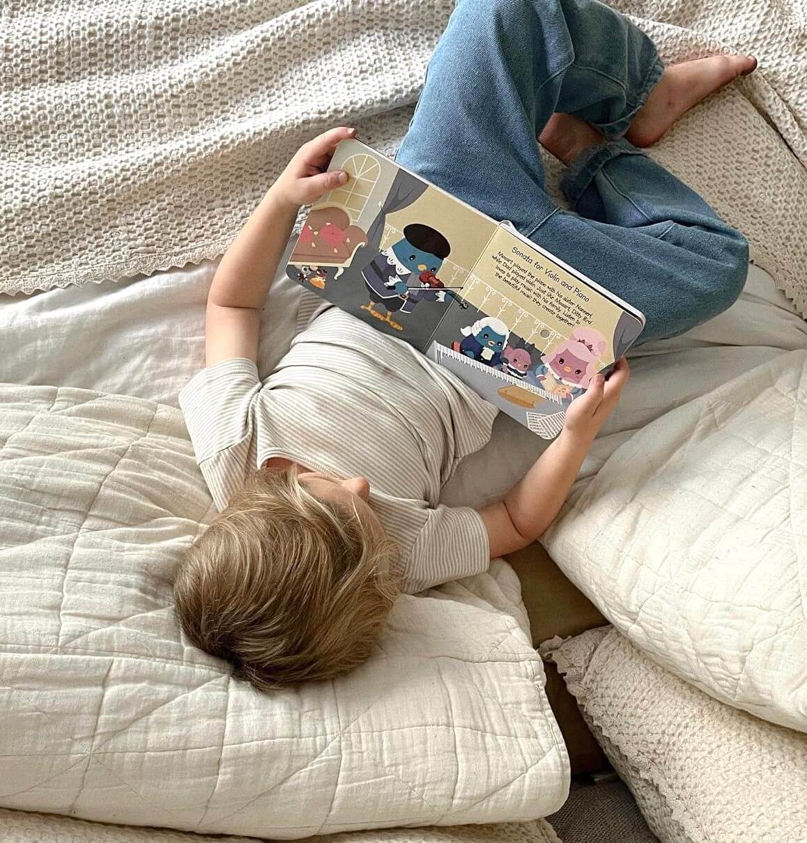 A young child lies on a white bed, reading Ditty Bird UK's "Music of Mozart"—a perfect educational book and first birthday gift that introduces little ones to classical music.