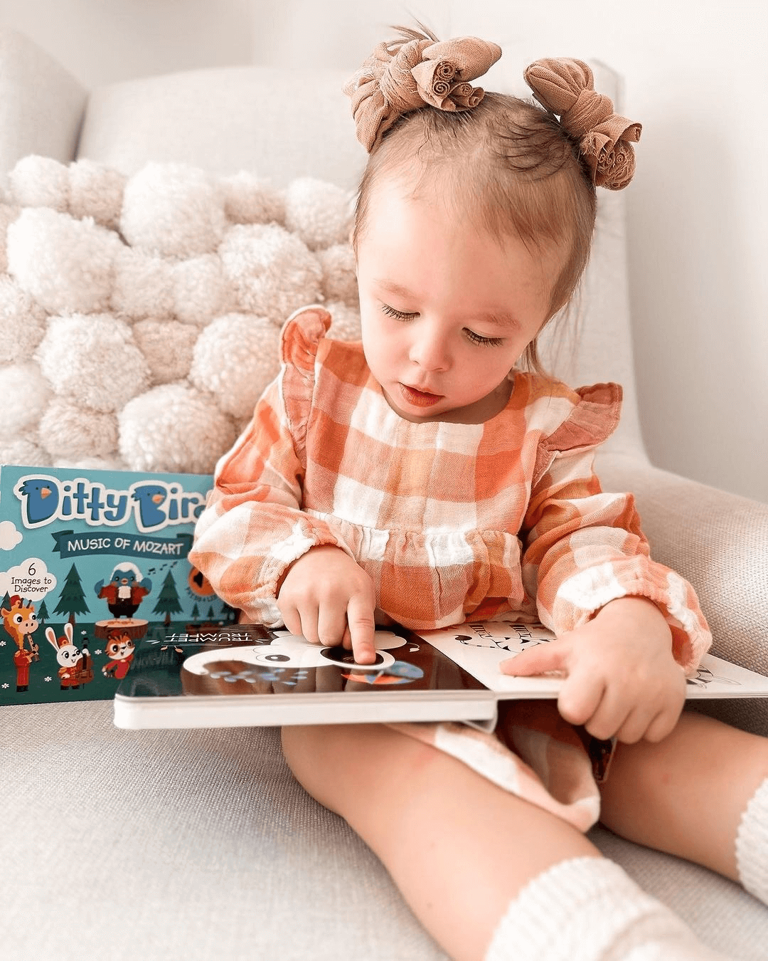 A young child in a peach plaid dress sits on a white chair, reading Ditty Bird UK's Music of Mozart—an interactive sound book that's a perfect first birthday gift or musical toy for toddlers aged 1-3.