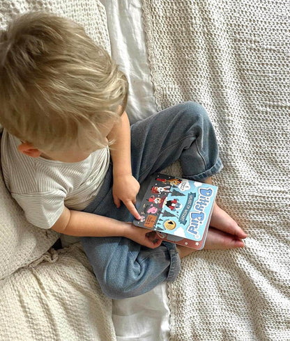 A young blonde-haired child sits on a bed with light-colored bedding, wearing a striped shirt and blue pants, exploring Ditty Bird UK’s "Music of Mozart" interactive sound book—an ideal first birthday gift.