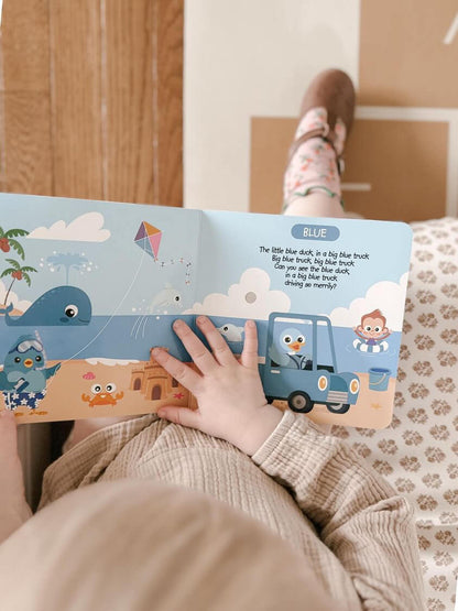 A child sits cross-legged, holding and reading Ditty Bird UK's colorful interactive sound book "Color Songs," featuring cartoon animals, a blue duck, blue truck, and a whale in a beach scene. Only the child's hand and legs are visible.