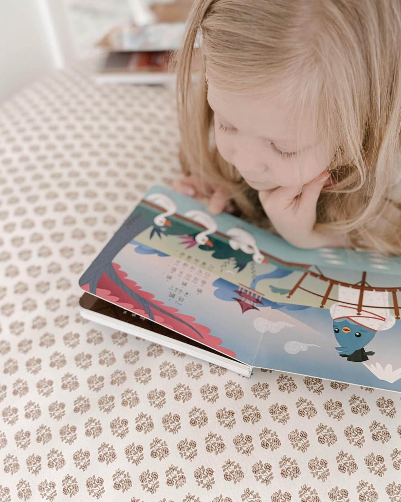 A young child with blonde hair lies on a patterned surface, exploring "Singing Chinese Poems" by Ditty Bird UK—an engaging book with colorful illustrations and music, perfect as an educational first birthday gift.