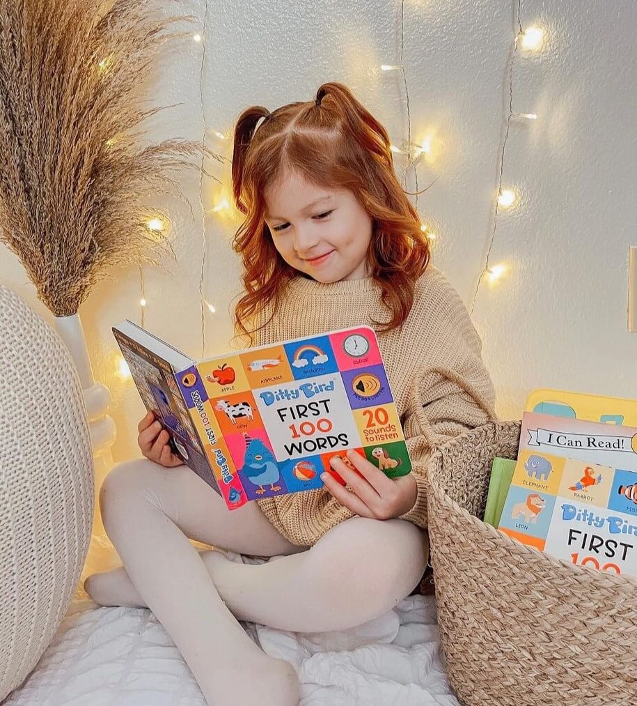 A young girl with brown hair in pigtails sits on her bed, smiling as she reads the Ditty Bird UK "100 Words Sound Book." Cozy decor and warm string lights create a perfect early learning atmosphere for exploring new words.