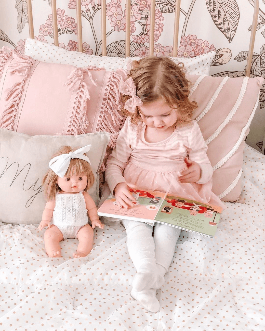A young girl in a pink dress sits on a bed with pink and white pillows, reading the "Funny Songs" interactive sound book by Ditty Bird UK, while a doll with a white bow and outfit rests beside her against floral wallpaper.
