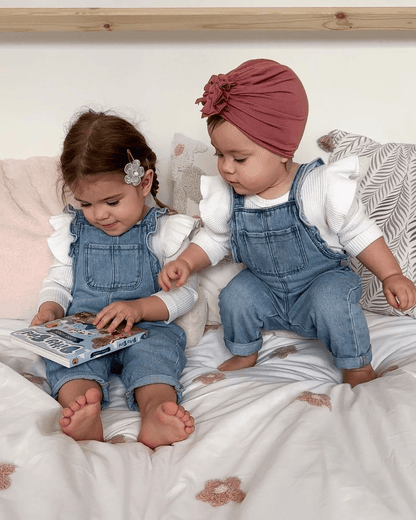 Two young children in matching denim overalls and white tops sit on a bed. One holds the Ditty Bird UK "Funny Songs" interactive sound book, while the other reaches for it. Both have brown hair—one with a pink headwrap, the other with a flower clip.