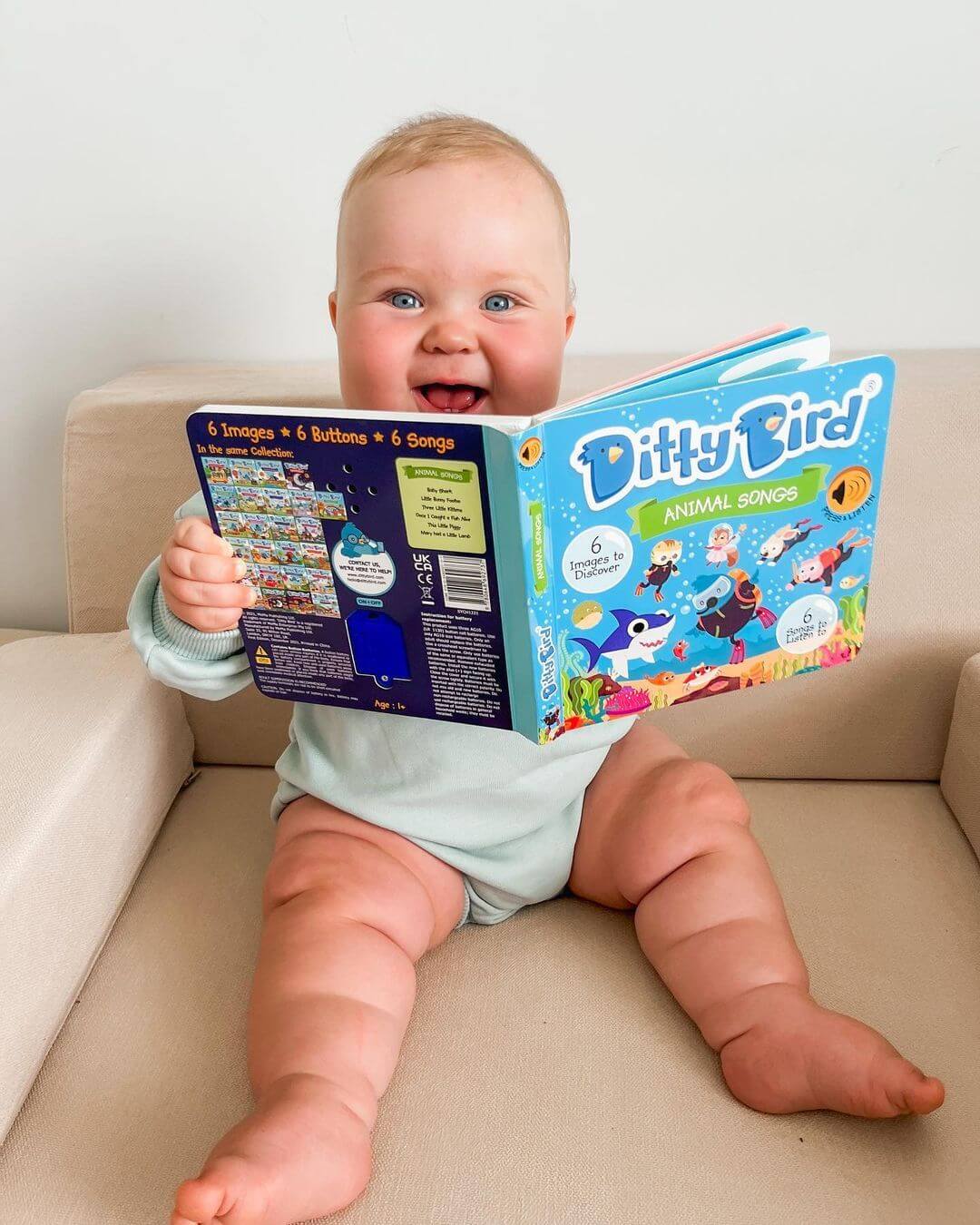 A smiling baby in a light blue onesie sits on a beige couch, holding and looking at an open “Ditty Bird UK Animal Songs” book—one of the best musical toys for toddlers aged 1-3 and a perfect first birthday gift.