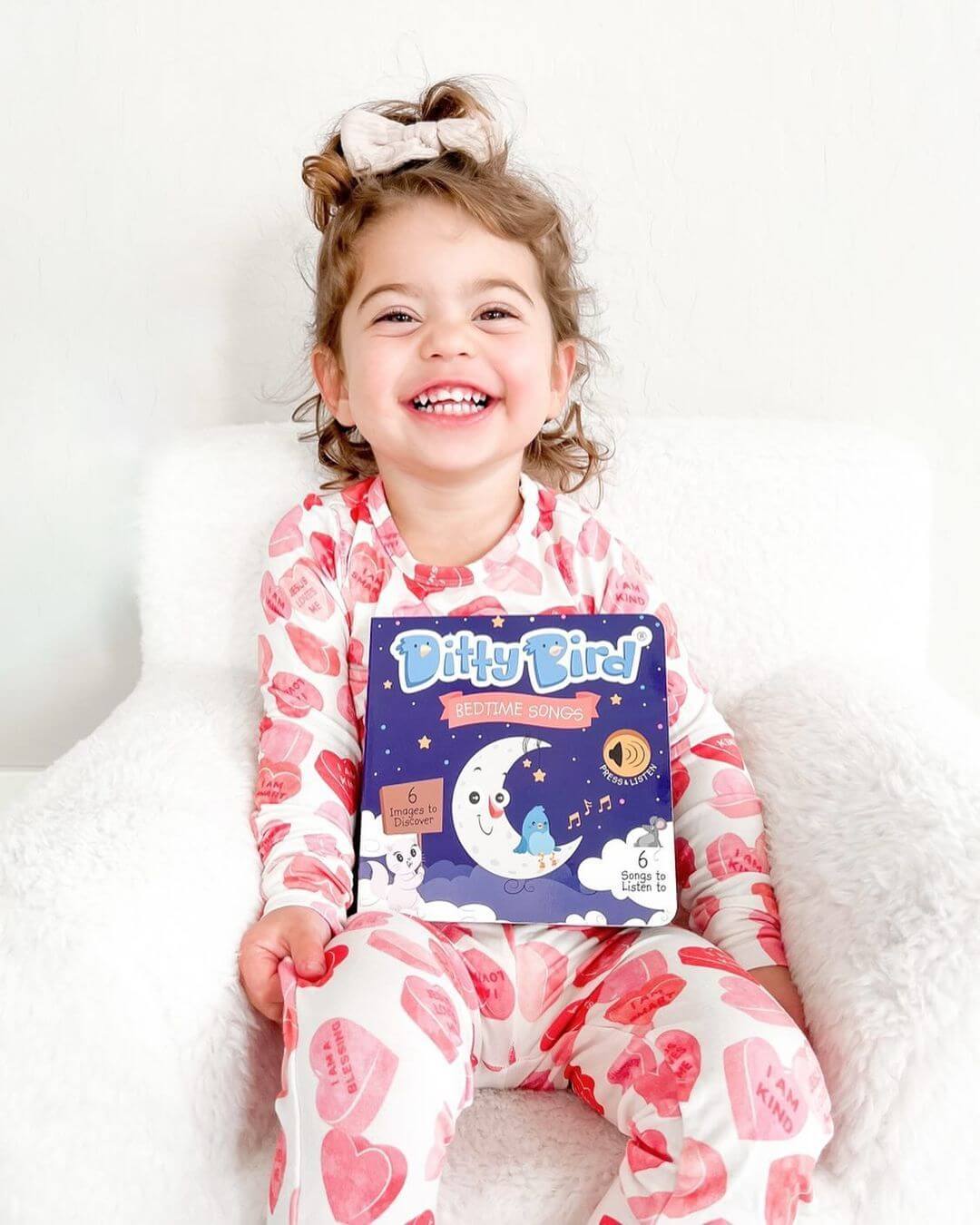 A young girl with curly hair, wearing heart-patterned pajamas, sits smiling on a white chair and holds Ditty Bird UK’s Bedtime Songs book—an interactive sound book and musical toy for toddlers ages 1-3.