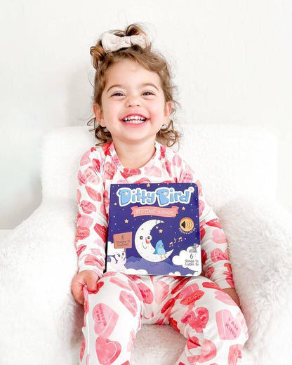 A young girl with curly hair, wearing heart-patterned pajamas, sits smiling on a white chair and holds Ditty Bird UK’s Bedtime Songs book—an interactive sound book and musical toy for toddlers ages 1-3.