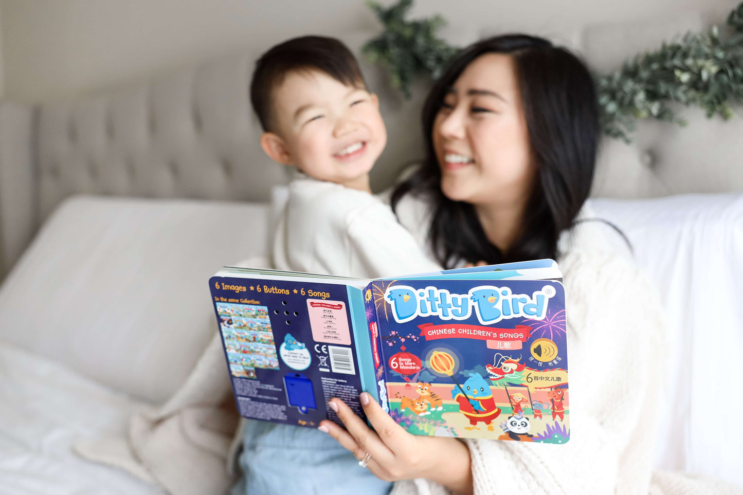 A smiling woman and a young boy sit on a bed reading Ditty Bird UK’s Chinese Children's Songs in Mandarin Vol.1—an engaging early learning book and perfect first birthday gift—with the colorful pages open towards the camera.