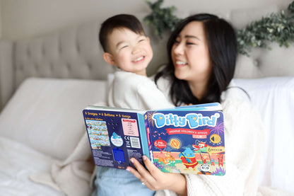 A smiling woman and a young boy sit on a bed reading Ditty Bird UK’s Chinese Children's Songs in Mandarin Vol.1—an engaging early learning book and perfect first birthday gift—with the colorful pages open towards the camera.
