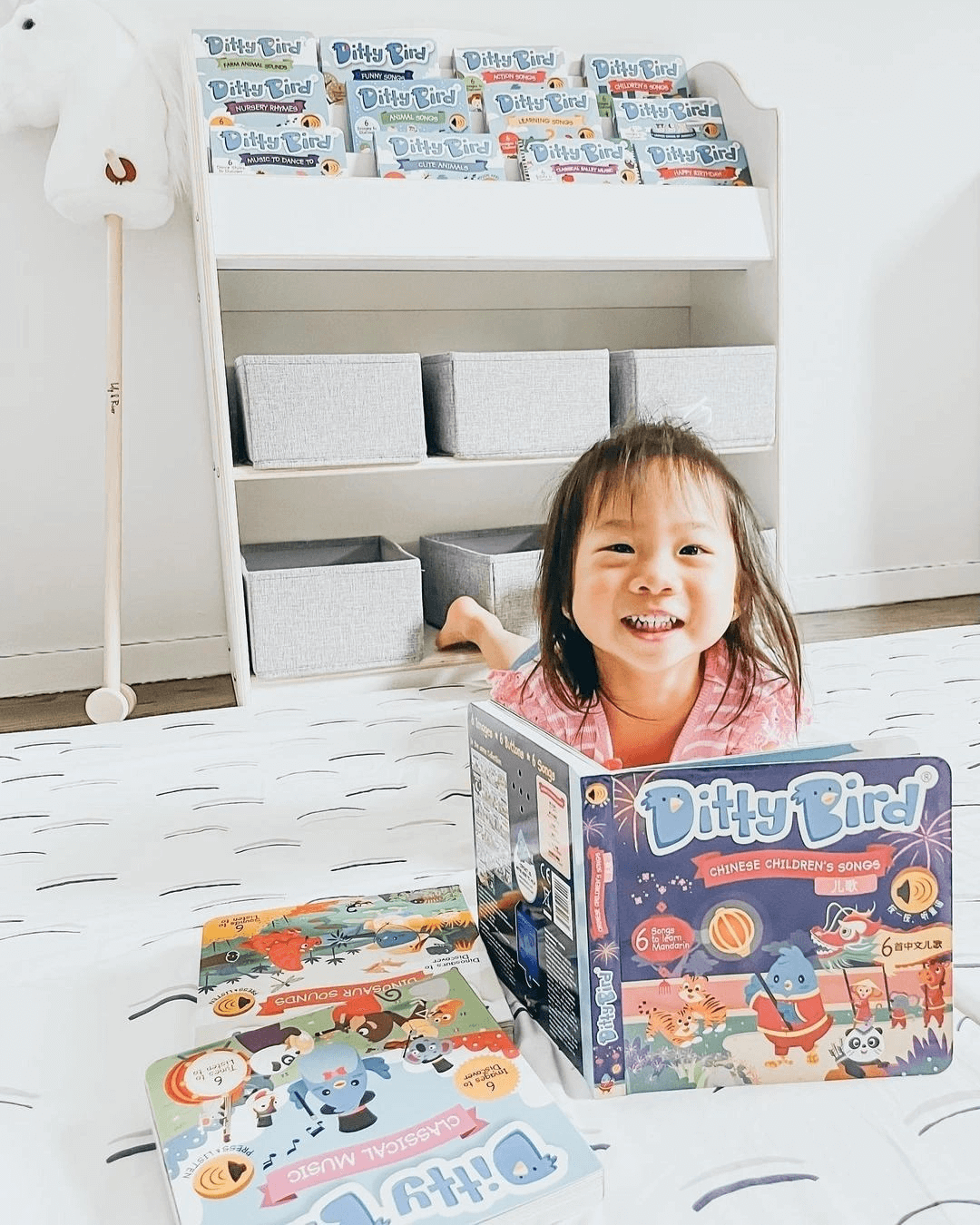 A smiling young girl lies on a bed holding Ditty Bird UK’s "Chinese Children's Songs in Mandarin Vol.1" interactive sound book. More Ditty Bird UK books are beside her, with a bookshelf and bins in the bright, child-friendly room.