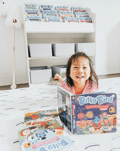 A smiling young girl lies on a bed holding Ditty Bird UK’s "Chinese Children's Songs in Mandarin Vol.1" interactive sound book. More Ditty Bird UK books are beside her, with a bookshelf and bins in the bright, child-friendly room.