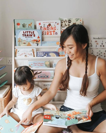 A woman with braided hair and a young girl sit on the floor, smiling as they read Ditty Bird UK's "Chinese Children's Songs in Mandarin Vol.2," surrounded by baby toys (6-12 months) and a bookshelf full of children's books.