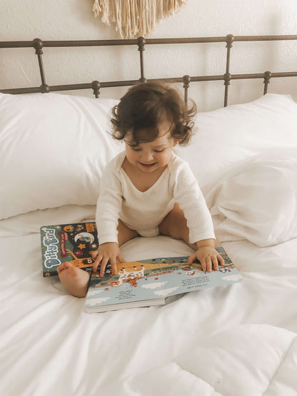 A smiling toddler with curly hair in a white onesie sits on a white bed, looking at the open Ditty Bird UK “Canciones de Animales en Español” book, with an educational toy and another closed book nearby for early learning fun.