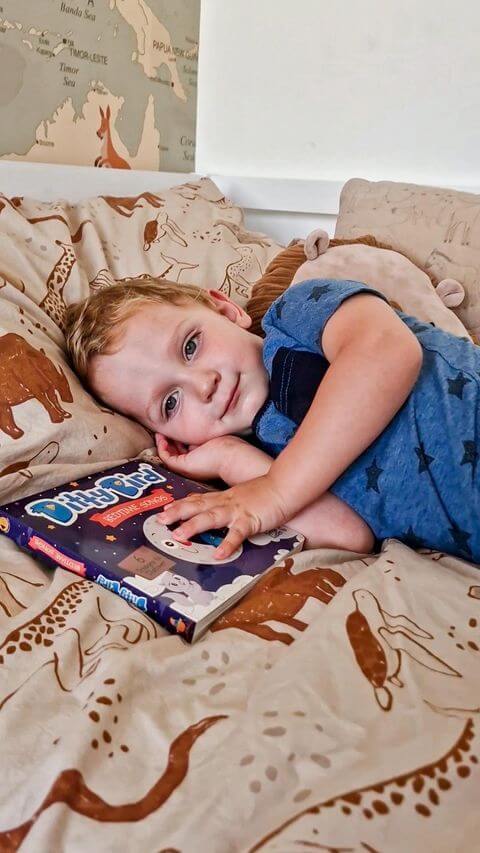 A young child with blonde hair lies on animal-themed bedding, cuddling Ditty Bird UK's interactive sound book, Bedtime Songs, and smiling softly at the camera. A world map is partially visible on the wall in the background.