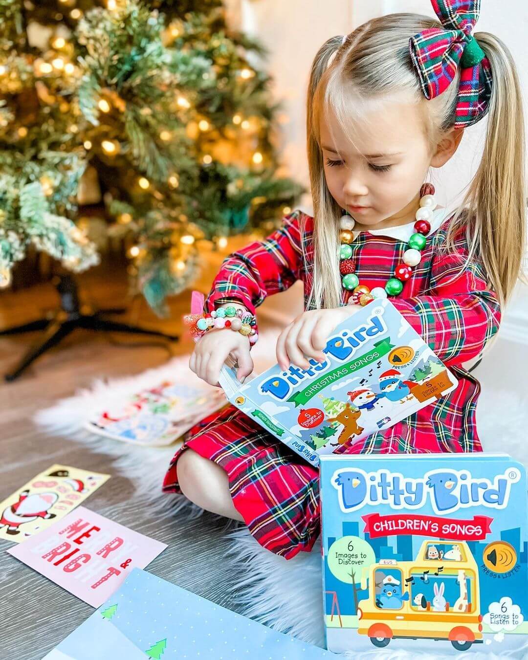 A young girl in a red plaid dress sits by the Christmas tree, opening a Ditty Bird UK “Children's Songs” book—one of the best musical toys for toddlers. Gifts and cards are scattered around her on the floor.