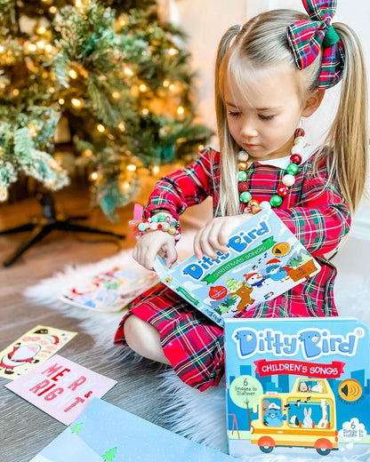 A young girl in a red plaid dress sits by the Christmas tree, opening a Ditty Bird UK “Children's Songs” book—one of the best musical toys for toddlers. Gifts and cards are scattered around her on the floor.