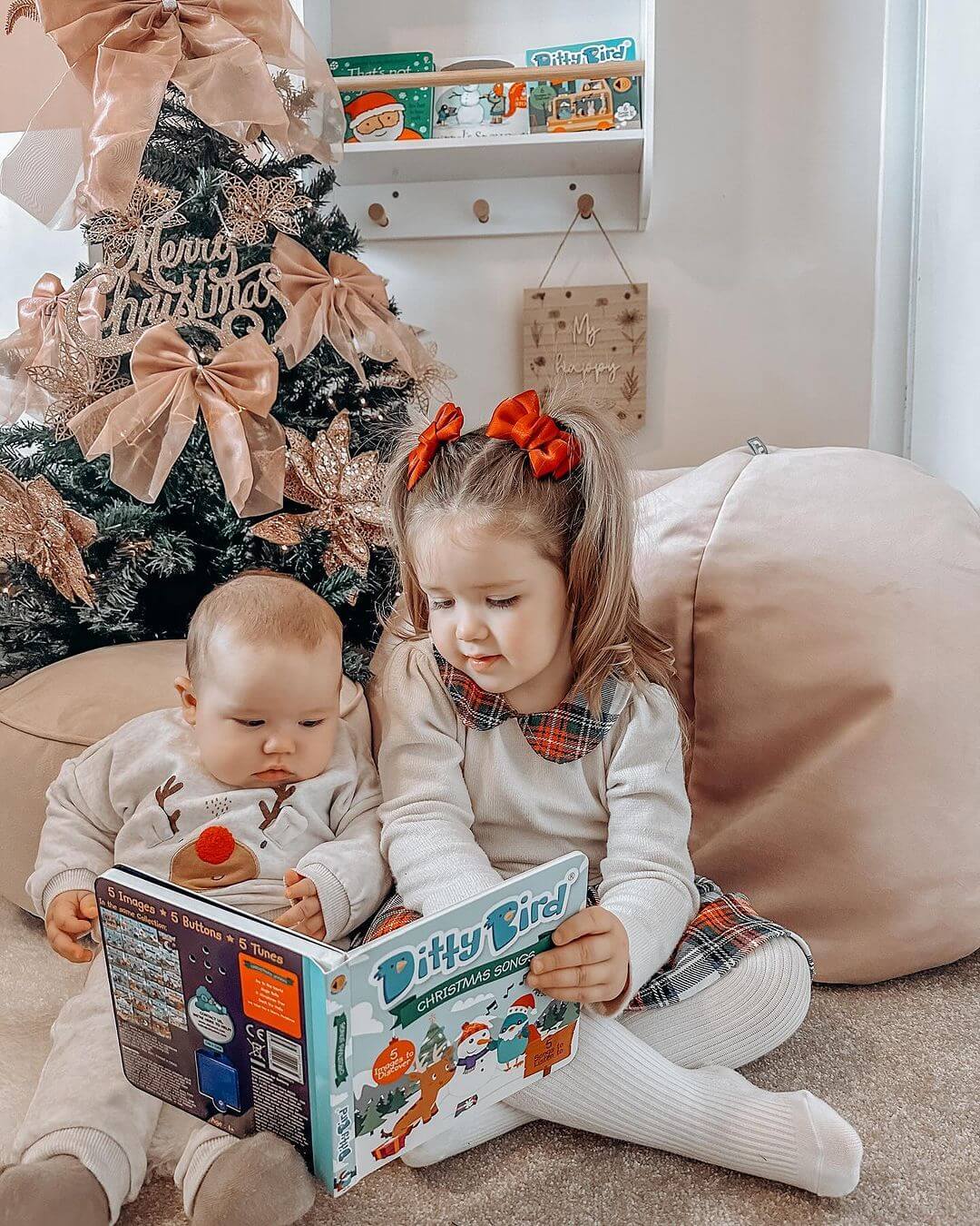 Two young children sit by the Christmas tree, reading "Christmas Songs" from Ditty Bird UK. The girl in a plaid dress with a red bow and the baby in white pajamas enjoy this festive moment with their interactive book.