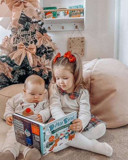Two young children sit by the Christmas tree, reading "Christmas Songs" from Ditty Bird UK. The girl in a plaid dress with a red bow and the baby in white pajamas enjoy this festive moment with their interactive book.