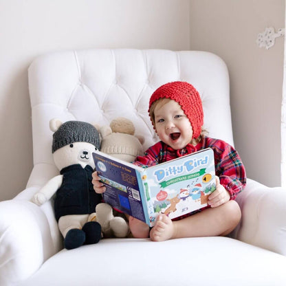 A smiling toddler in a red knit hat and plaid outfit sits on a white armchair, holding an open Ditty Bird UK "Christmas Songs" book—an ideal educational toy—with two stuffed bears beside them.