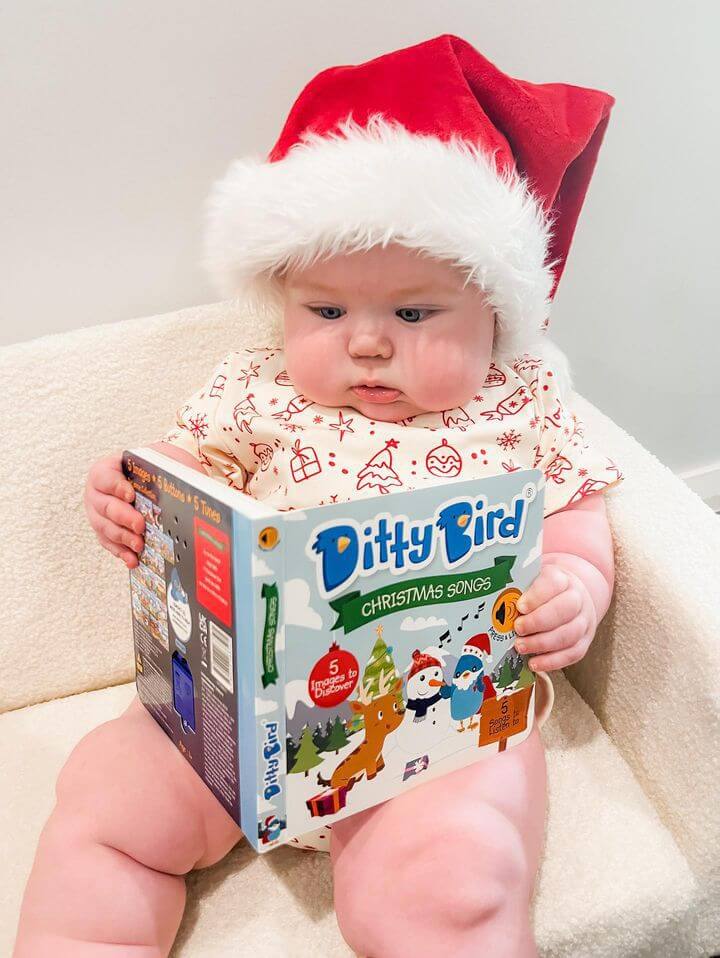 A baby in a Santa hat and festive pajamas sits on a chair, holding and looking at the Ditty Bird UK "Christmas Songs" book—an ideal first birthday gift and one of the best musical toys for toddlers ages 1-3.