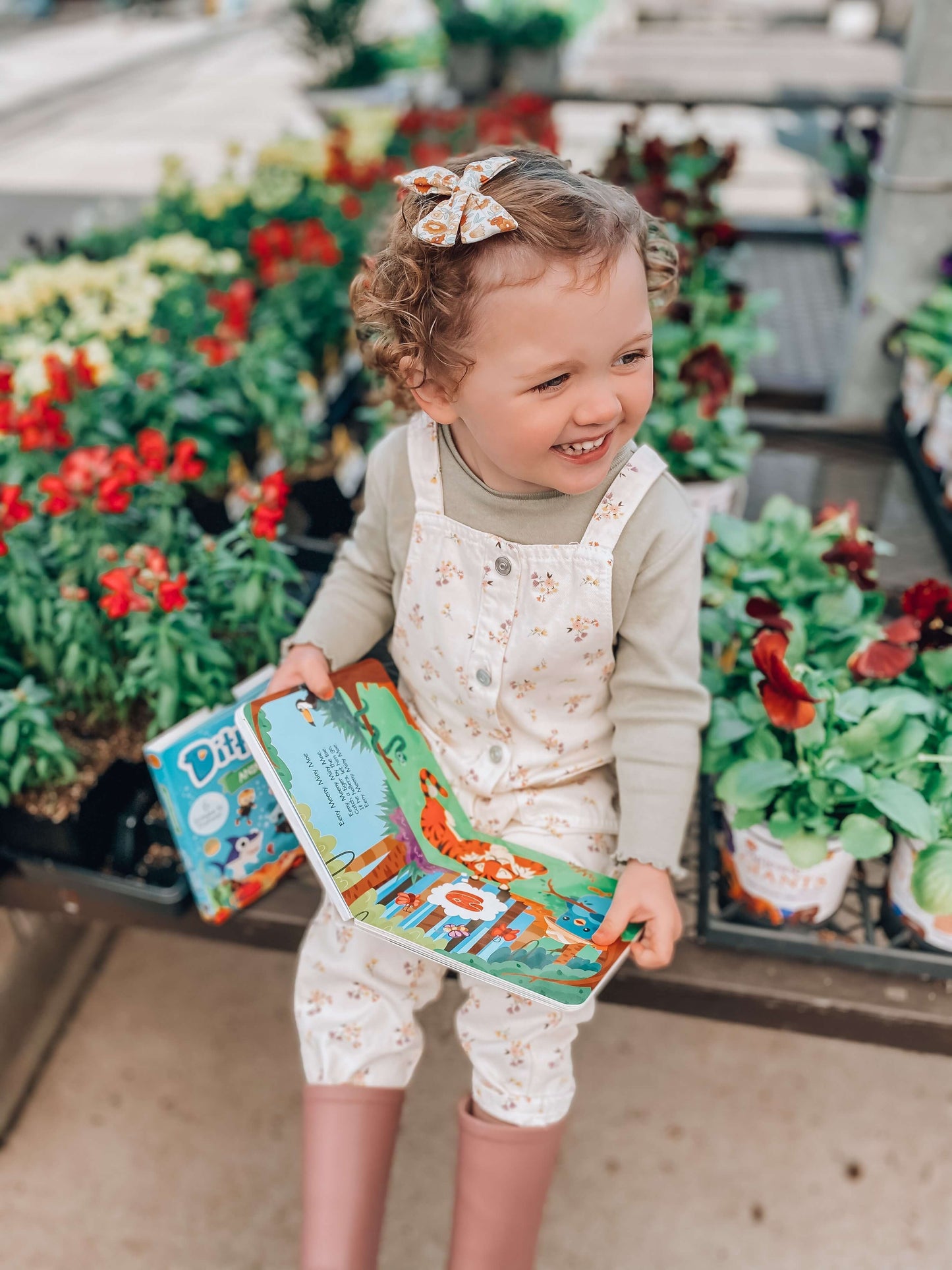 A young child with curly hair and a floral bow sits among potted plants, smiling and holding Ditty Bird UK’s "Funny Songs" interactive sound book—an ideal first birthday gift—wearing a light outfit and pink rain boots.