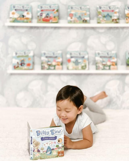 A young child smiles while lying on a white bed, reading the Ditty Bird UK "Funny Songs" book—an ideal educational toy. Behind them, more Ditty Bird UK books are neatly arranged on two white shelves against a patterned wall.